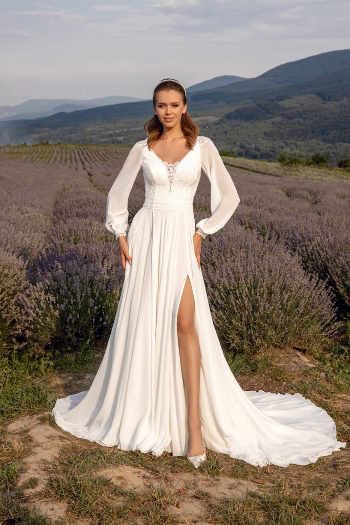 In questa foto una modella posa indossando un abito da sposa scivolato in chiffon con spacco e maniche lunghe a sbuffo di Angela Bianca