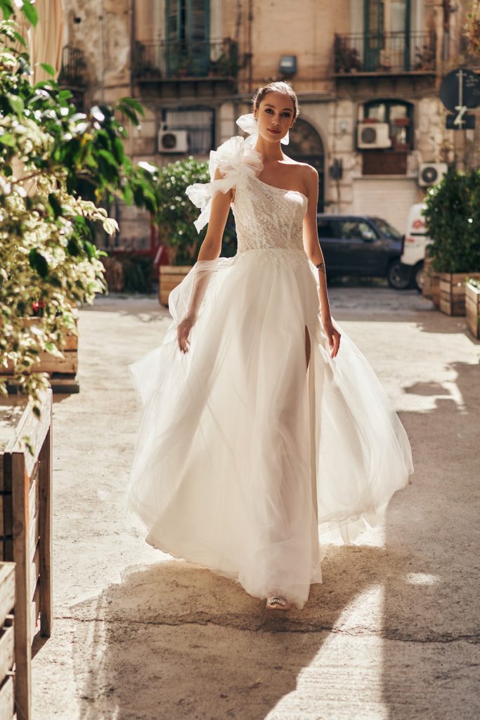 In questa foto una modella posa indossando un abito da sposa ampio in organza di seta, monospalle con maxi fiore sulla spalla destra di Annai Bridal