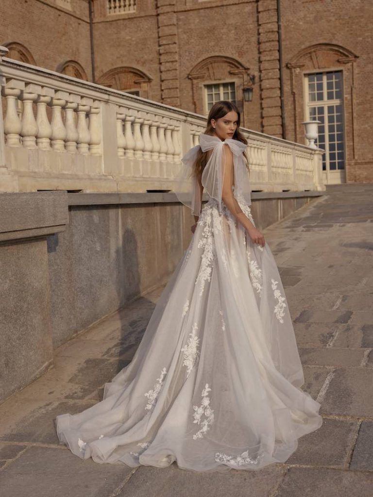 In questa foto una modella posa indossando un abito da sposa di Capri Sposa in tulle, con maxi fiocchi sulle spalle e decorazioni floreali sulle lunghezze