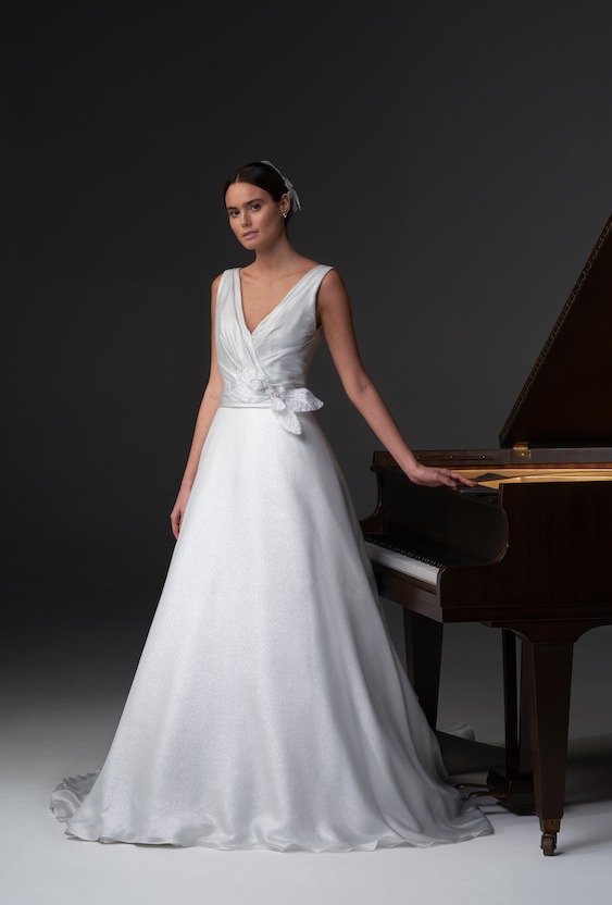 In questa foto una modella posa indossando un abito da sposa ad A in organza di seta con scollo a V e un fiore 3D su un fianco firmato da Chiara Vitale - Atelier Kore