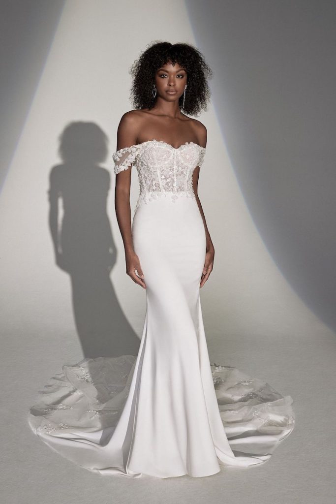 In questa foto una modella posa indossando un abito da sposa a semi sirena in chiffon con bustier in pizzo, scollo a cuore e manichette di Justin Alexander