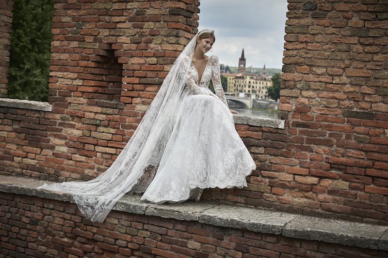 In questa foto una modella posa a Verona indossando un abito di Maison  Signore. L'abito è ampio, in pizzo chantilly, con maniche lunghe e scollo a V. La modella indossa anche un velo abbinato in pizzo