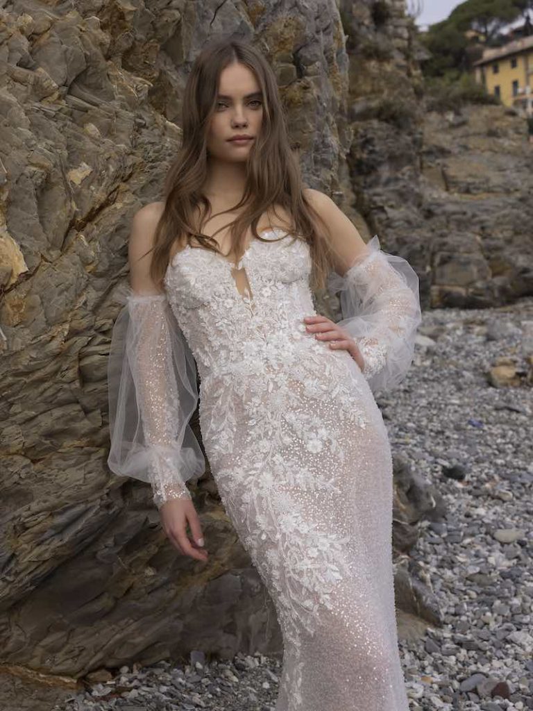 In questa foto una modella posa indossando un abito da sposa a sirena di Michela Ferriero con scollo a cuore, maniche in tulle e cristalli removibili, fiori 3D e cristalli su corpetto che scendono ramage sulle lunghezze