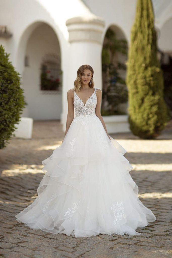 In questa foto una modella posa indossando un abito da sposa principessa con scollo a V e tulle a balze di Monica Loretti