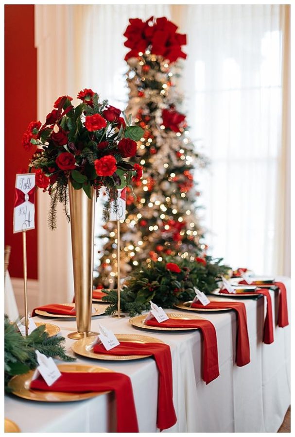 In questa foto una tavola per matrimonio a tema Natale decorata con piatti di colore oro su cui sono poggiati tovaglioli di colore rosso, runner di rami di pino e rose di colore rosso, un'alzatina con rose di colore rosso. Sullo sfondo si intravede un albero di Natale decorato
