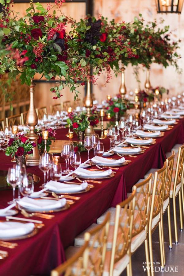 In questa foto un tavolo per matrimonio a tema Natale con tovaglia di colore bordeaux e tovaglioli di colore bianco. Il tavolo è decorato con piccole composizioni di rose di colore rosso e alzatine di colore oro con composizioni floreali abbinate. Accanto al tavolo sono presenti sedie chiavarine di colore oro 