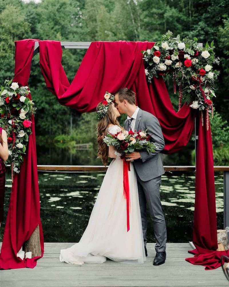 In questa foto due sposi si abbracciano e baciano davanti ad un arco di legno decorato con drappi di colore rosso, foglie e rose di colore bianco e rosso. La sposa ha tra i capelli una corona di fiori e porta nella mano destra un bouquet abbinato