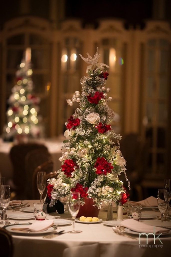 In questa foto un centrotavola per matrimonio a tema Natale composto da un piccolo albero decorato con fiori e fiocchi di colore rosso, fiori di colore bianco, palline di colore argento