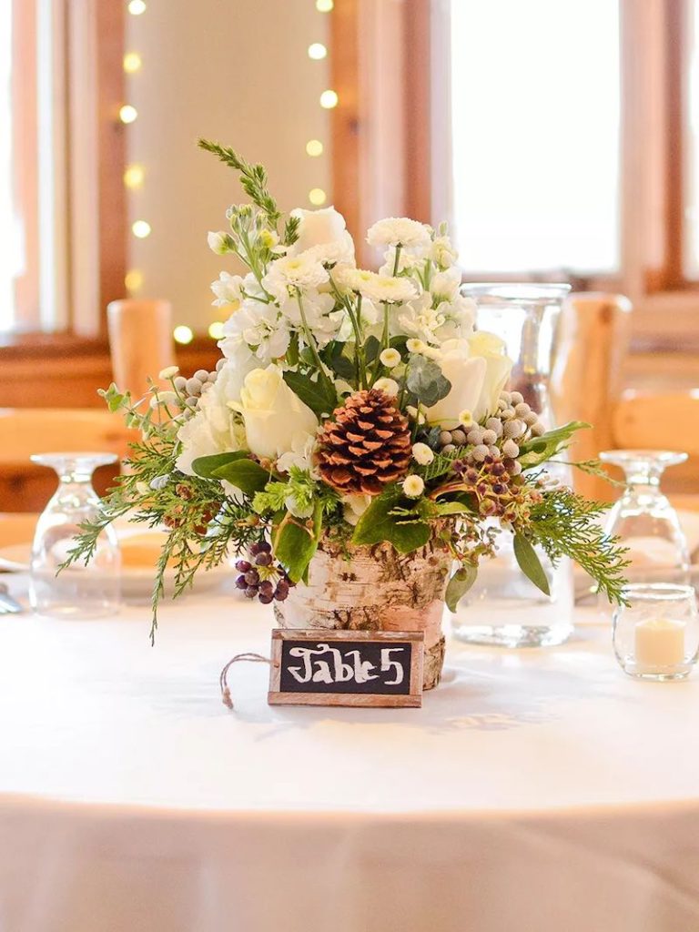 In questa foto un centrotavola per matrimonio a tema Natale composto da fiori ci colore bianco, aghi di pino e pigne 