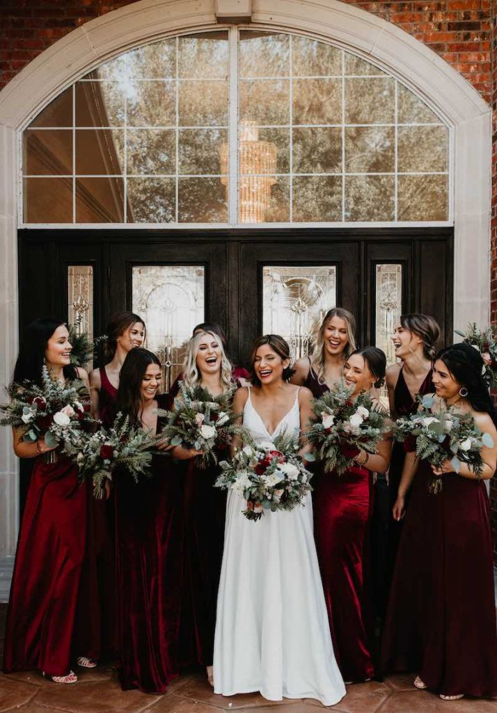 In questa foto una sposa ride circondata dalle sue damigelle che indossano abiti di velluto di colore rosso e tengono tutte tra le mani bouquet di rami di pino, eucalipto e fiori di colore rosso e bianco