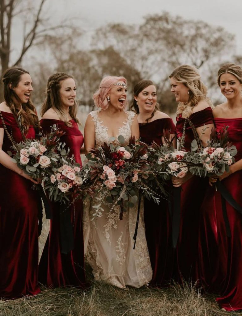 In questa foto una sposa a Natale circondata dalle sue damigelle che indossano un abito di velluto di colore rosso, ridono e tengono tra le mani bouquet di pino e rose di colore rosso e rosa