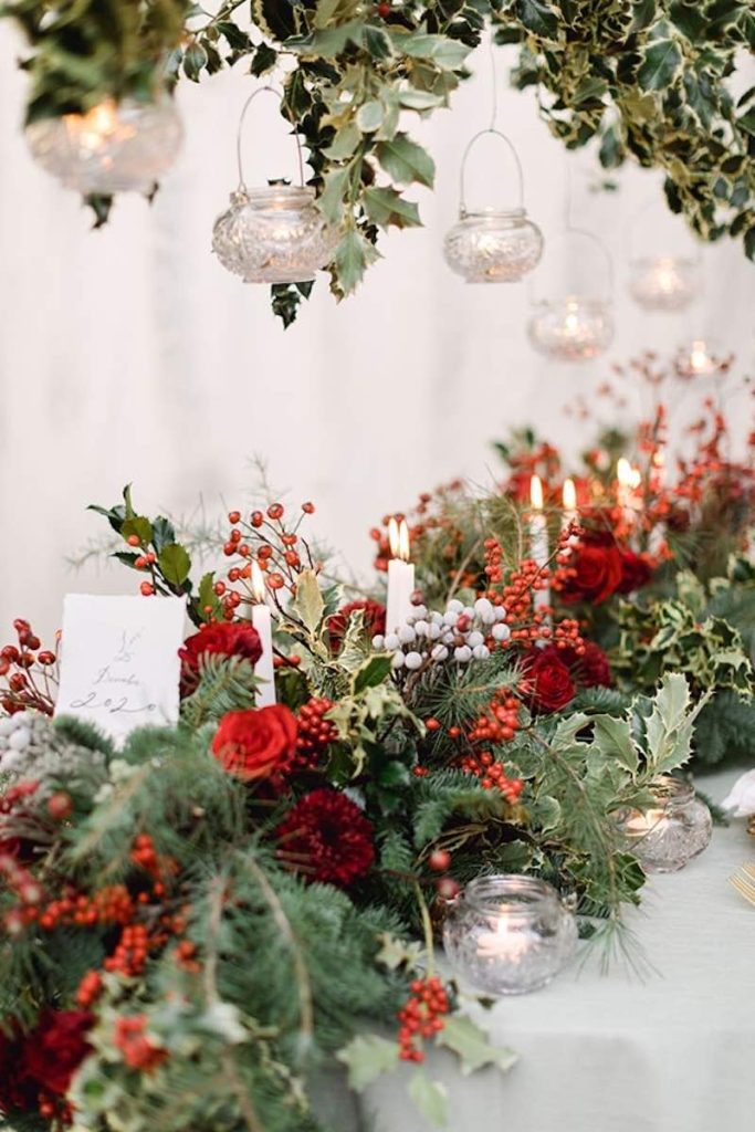 In questa foto una composizione di fiori per matrimonio natalizio con rami di pino, rose di colore rosso, bacche di colore rosso e bianco decorata con candele di colore bianco