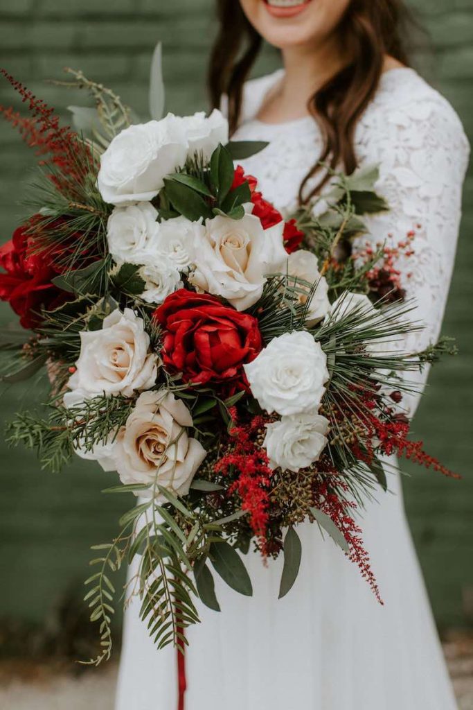 In questa foto una sposa tiene in mano un bouquet a tema Natale con rami di pino, bacche di colore rosso, rose di colore bianco e rosso