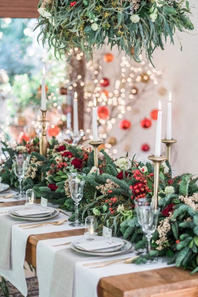 In questa foto il dettaglio di una tavola per matrimonio a tema Natale con runner di rami di pino, bacche di colore rosso e spighe decorato portacandele di colore oro e candele di colore bianco. Il tavolo è sovrastato da una composizione abbinata al runenri. 