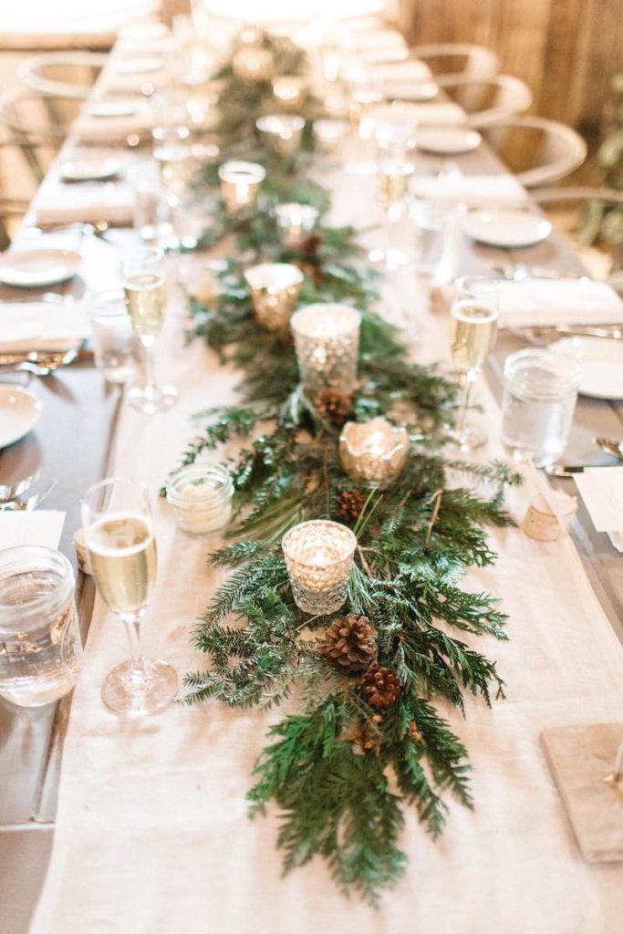 In questa un tavolo imperiale per matrimonio a tema Natale decoarato con un runner di rami di pino con pigne e portacandele bassi di vetro 