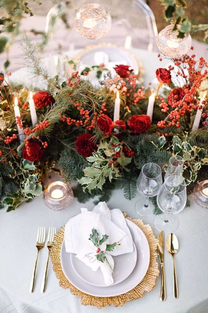 In questa foto il dettaglio di una mise en place di un matrimonio natalizio composta da un sottopiatto di colore oro con posate abbinate e un runner di rami di pino, agrifoglio, bacche, rose di colore rosso e candele accese di colore bianco. Sopra il piatto è presente un tovagliolo di colore bianco fermato da un segnaposto con agrifoglio