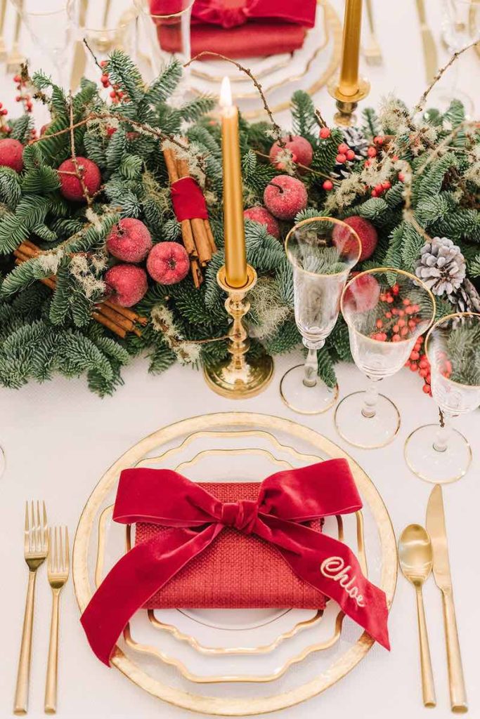 In questa foto il dettaglio di una mise en place per matrimonio a tema Natale con segnaposto a forma di fiocco velluto di colore rosso con il nome di un ospite poggiato su piatti di porcellana bianca e bordo di colore oro abbinati a posate di colore oro. Il tavolo è decorato da un runner di rami di pino, frutta di colore rosso, bacche e pigne