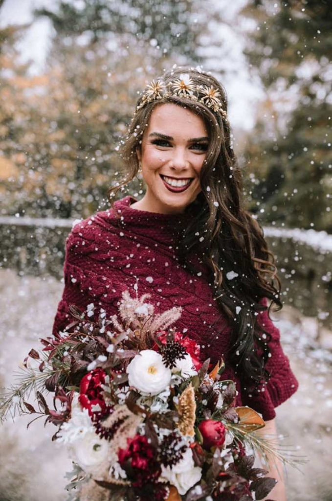 In questa foto una sposa indossa una stola di lana di colore rosso e tiene tra le mani un bouquet di fiori colore bianco e rosso. La sposa porta tra i capelli un cerchietto con stelle alpine gioiello