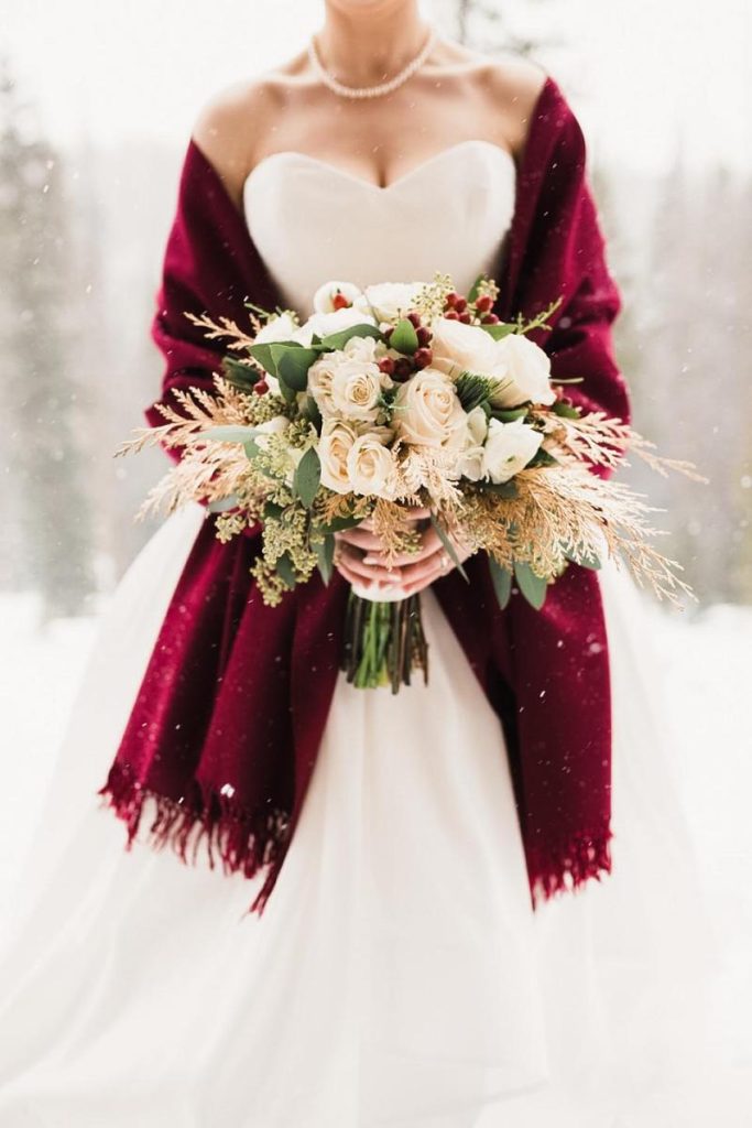 In questa foto una sposa a Natale indossa una stola di lana di colore rosso e tiene tra le mani un bouquet di rose di colore bianco e spighe