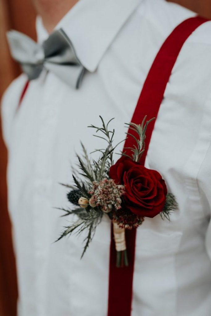 In questa foto il dettaglio del fiore all'occhiello di uno sposo con piccole pigne e una rosa di colore rosso attaccato ad una bretella di colore rosso