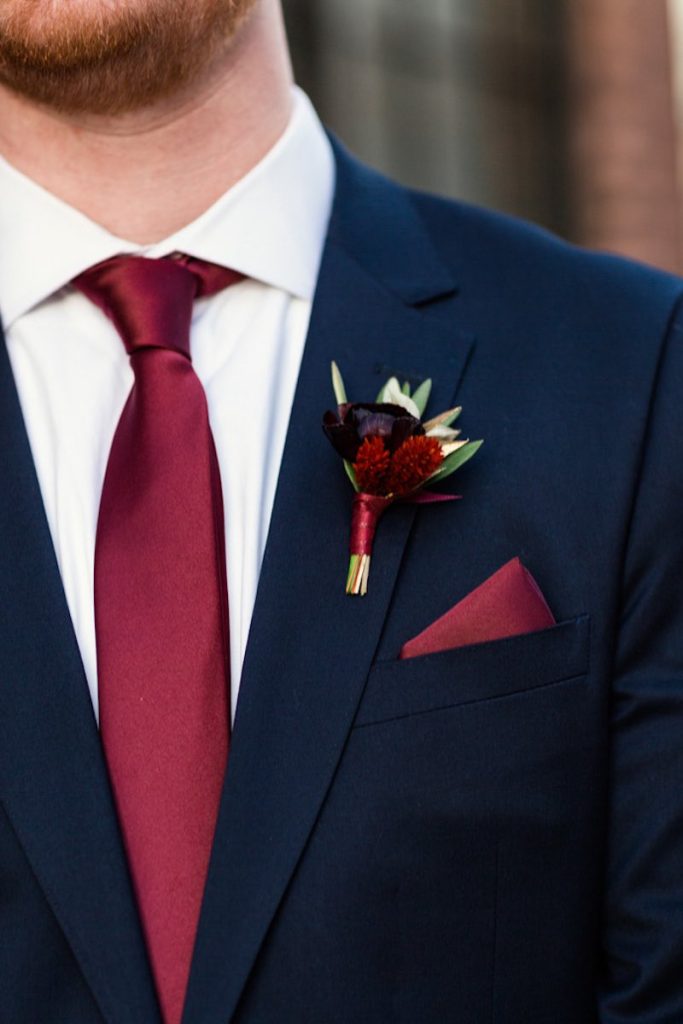 In questa foto il dettaglio di un look da sposo con cravatta di colore borgogna, fazzoletto nel taschino abbinato e fiore all'occhiello con fogli e fiori di colore rosso e bordeaux