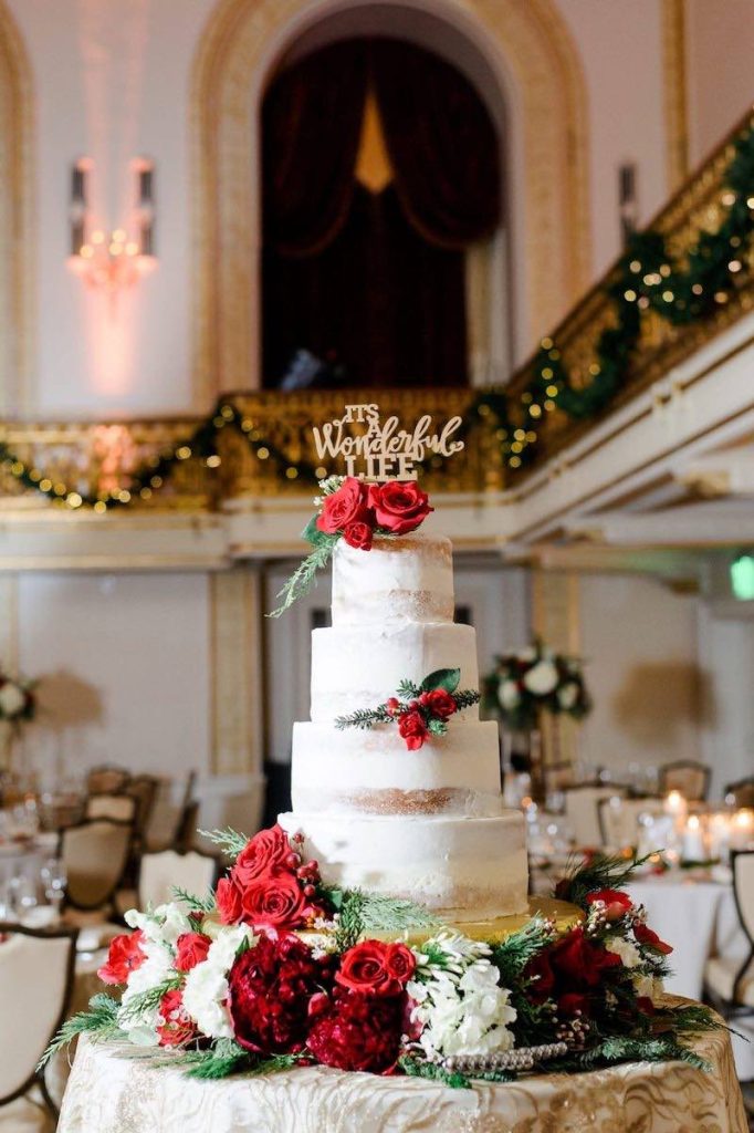 In questa foto una torta per matrimonio a tema Natale di colore bianco decorata con fiori di color rosso e un cake topper di colore dorato