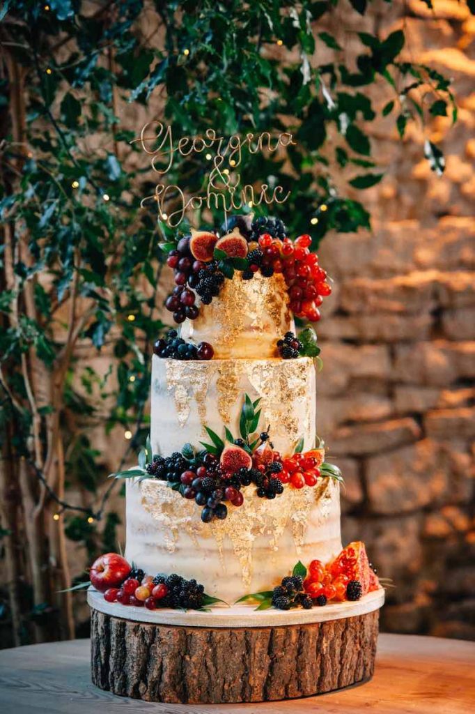 In questa foto una torta per matrimonio decorata con foglia oro, frutti rossi e un cake topper dorato con i nomi degli sposi