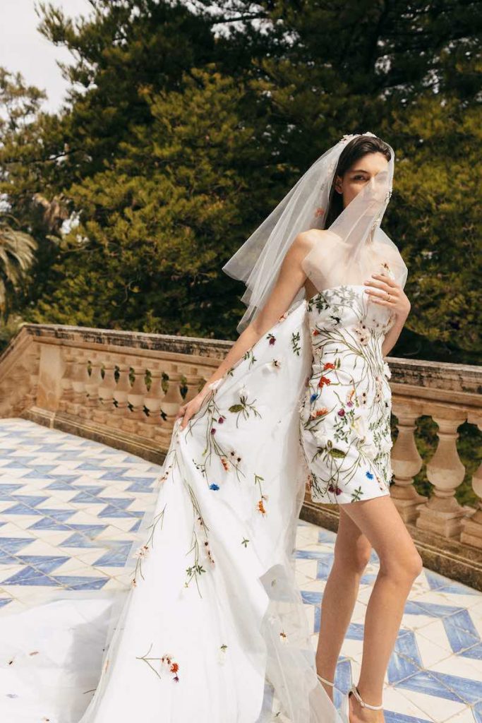 In questa foto una modella indossa un abito da sposa corto con lungo strascico con ricami floreali 3D multicolor di Monique Lhuillier da usare come secondo abito da sposa