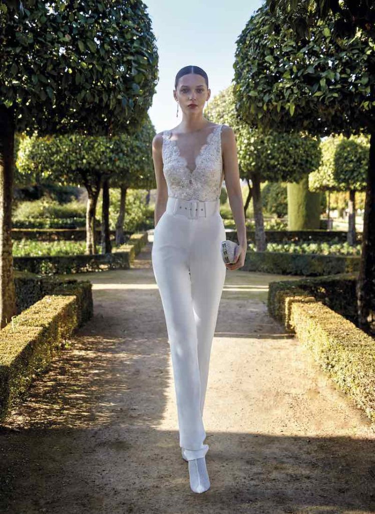 In questa foto una modella indossa una tuta da sposa con pantaloni aderenti, bustier di pizzo con scollo a V e cinturone in vita di Valerio Luna