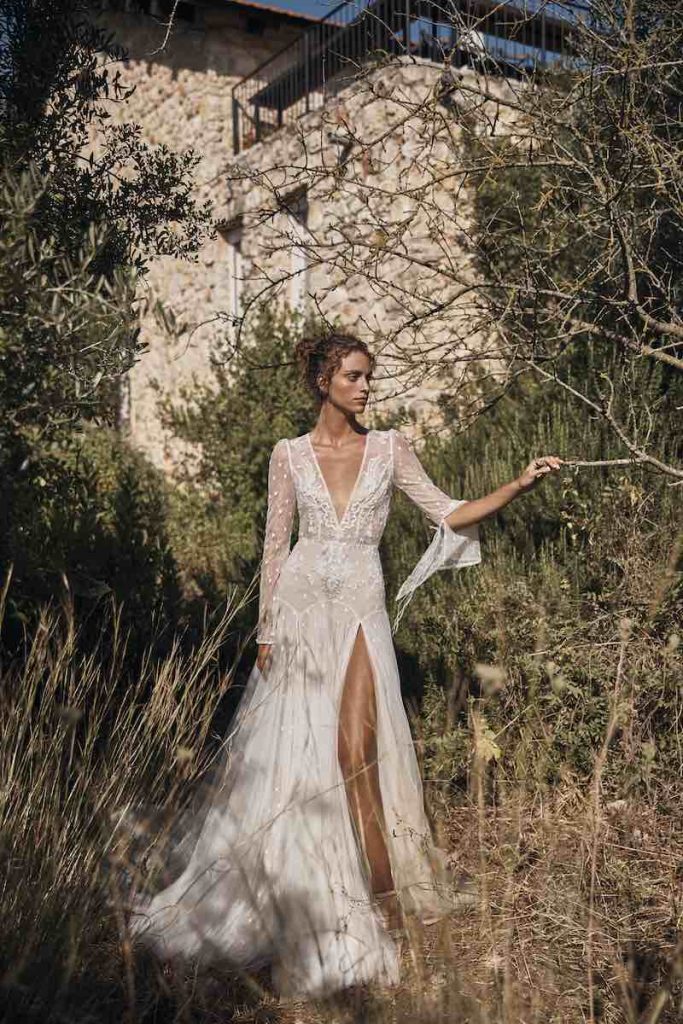 In questa foto una modella indossa un abito da sposa con spacco, scollo a V e maniche ampie di Gali Karten