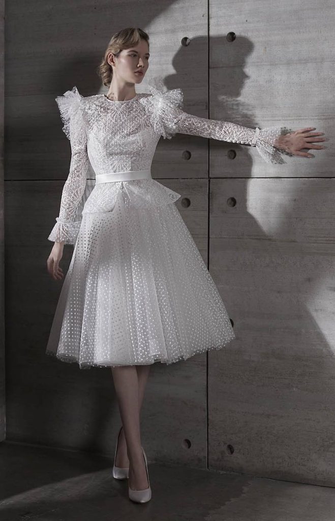 In questa foto una modella indossa un abito da sposa corto con gonna a ruota, maniche e ruches sulle spalle in tulle plumetis di Elisabetta Polignano