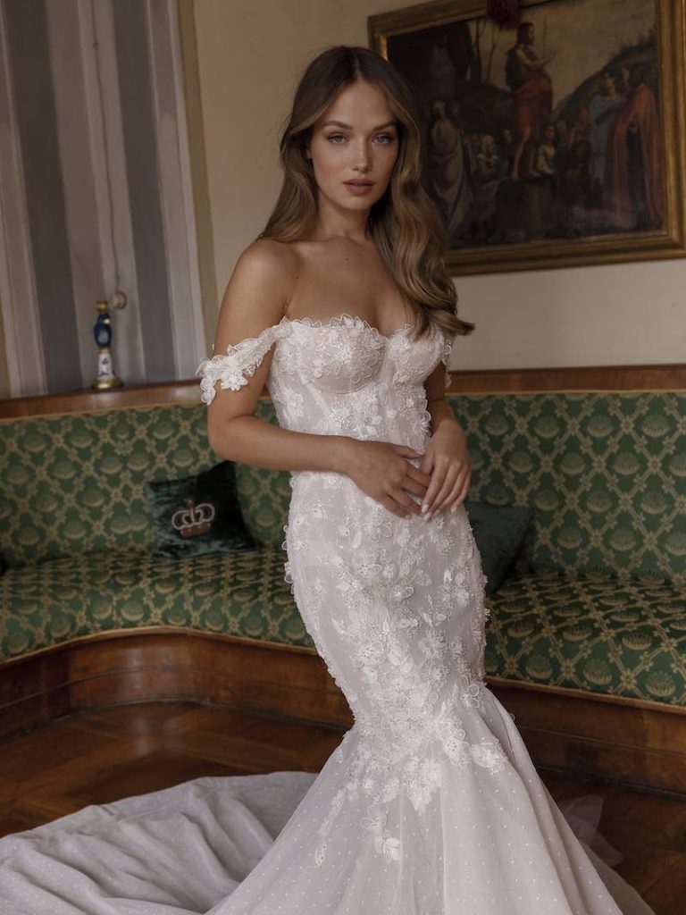 In questa foto una modella posa con un abito da sposa semisirena di Ricca Sposa, con maniche scivolate e applicazioni 3D all over