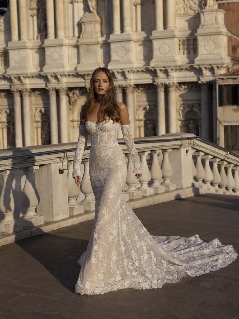 In questa foto una modella passeggia indossando un abito da sposa a sirena, con bustier e maniche rimovibili della collezione Ricca Sposa 2023