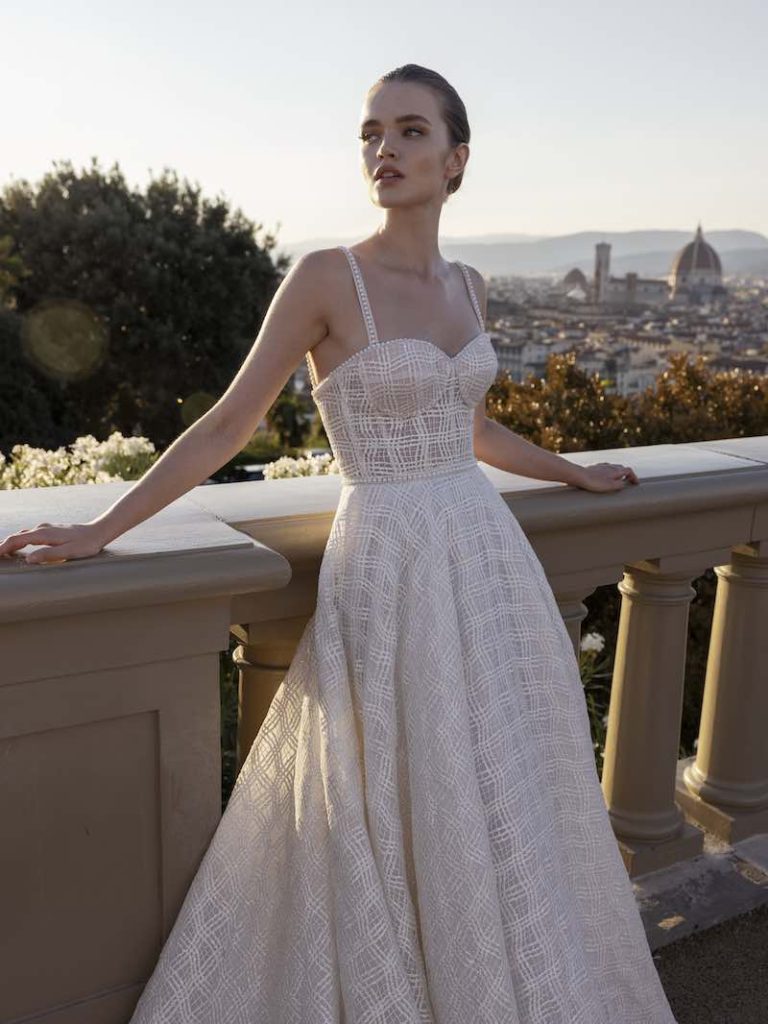 In questa foto una modella indossa un abito da sposa con gonna scivolata e corsetto. Le bretelle sono sottilissime. Di pregio la lavorazione del tessuto, che sembra ricreare un motivo a quadri ton sur ton