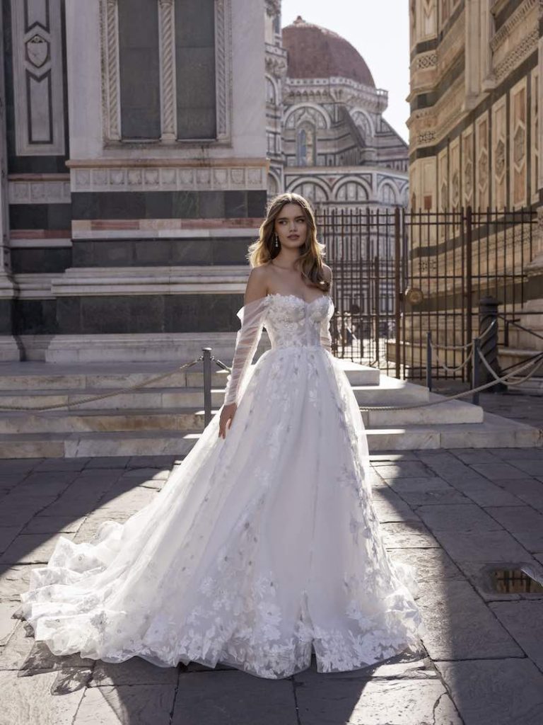 In questa foto una modella, a Firenze, indossa un abito da sposa con bustier e gonna scivolata con ricami in pizzo della collezione Ricca Sposa 2023. Le maniche sono in tulle, lunghe e scivolate