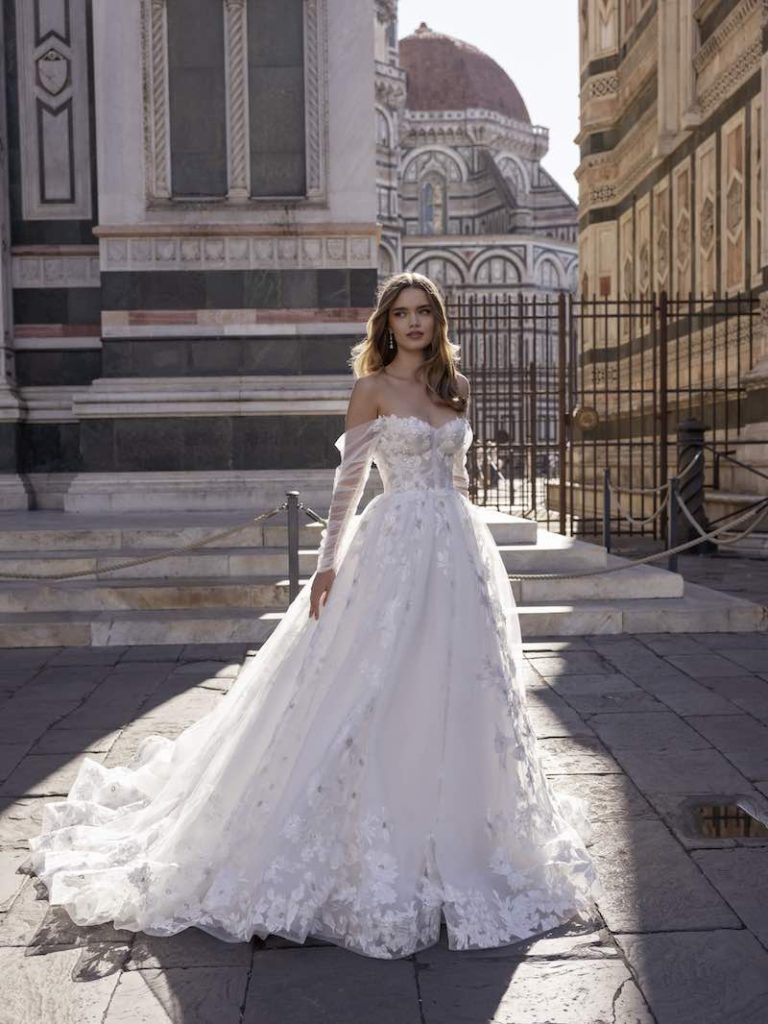 In questa foto una modella, a Firenze, indossa un abito da sposa con bustier e gonna scivolata con ricami in pizzo della collezione Ricca Sposa 2023. Le maniche sono in tulle, lunghe e scivolate