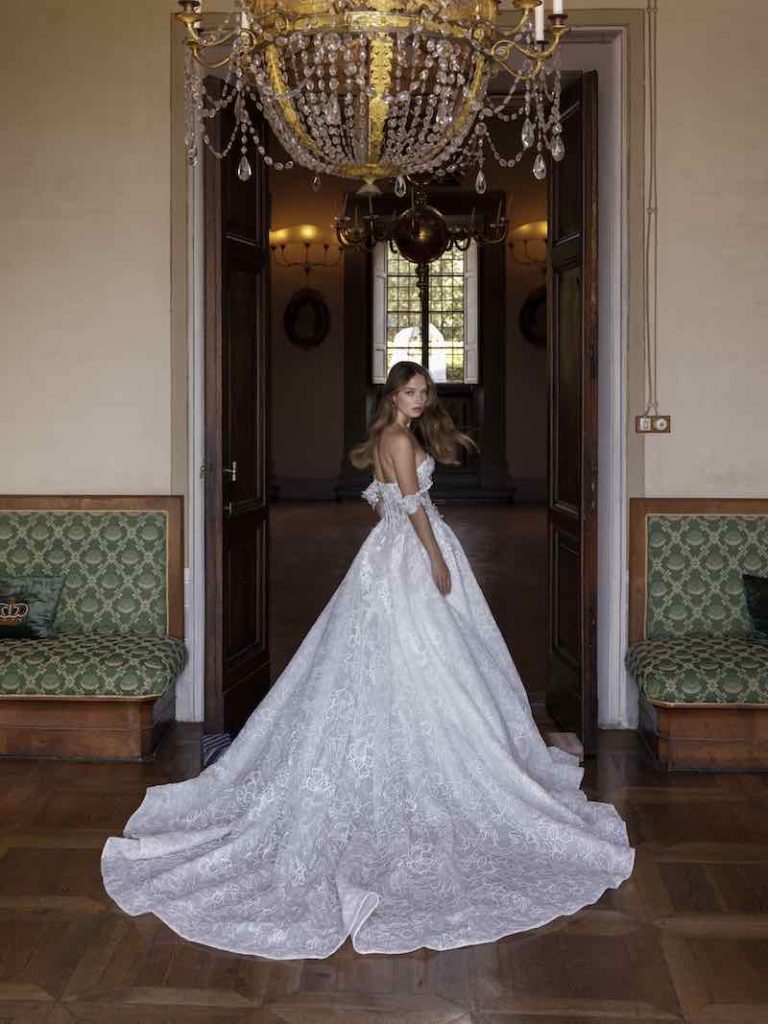 In questa foto, una modella di spalle all'interno di un grande salone mentre indossa un abito da sposa dalla linea ad A di Ricca Sposa 2023