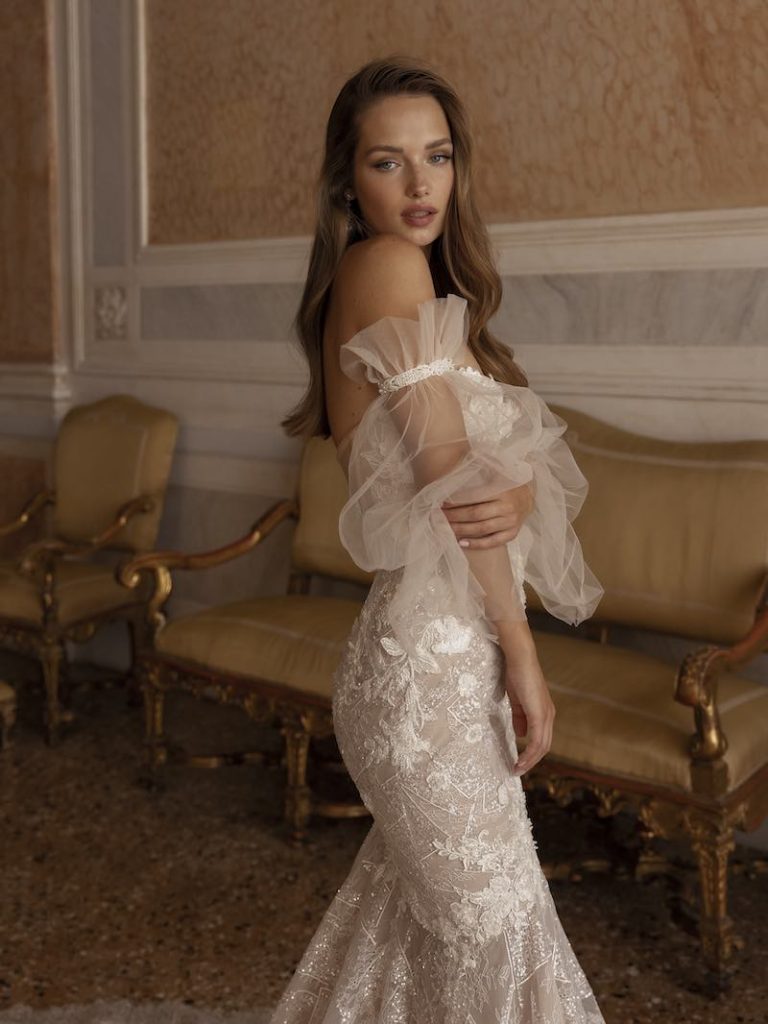 In questa foto una modella posa con un abito da sposa sirena 2023 della collezione La mia Italia di Ricca Sposa. Le maniche balloon, in tulle, lasciano le spalle scoperte. Sono rimovibili. 
