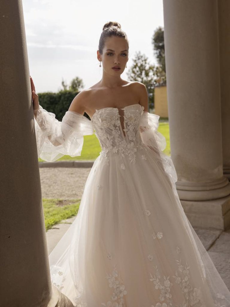 In questa foto una modella indossa un abito da sposa con maniche lunghe e scivolate. Sul corpetto sono applicati fiori 3D