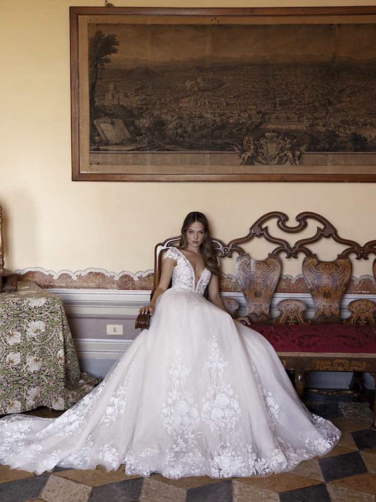In questa foto una modella seduta indossa un abito da sposa scivolato e con profonda scollatura a V di Ricca Sposa