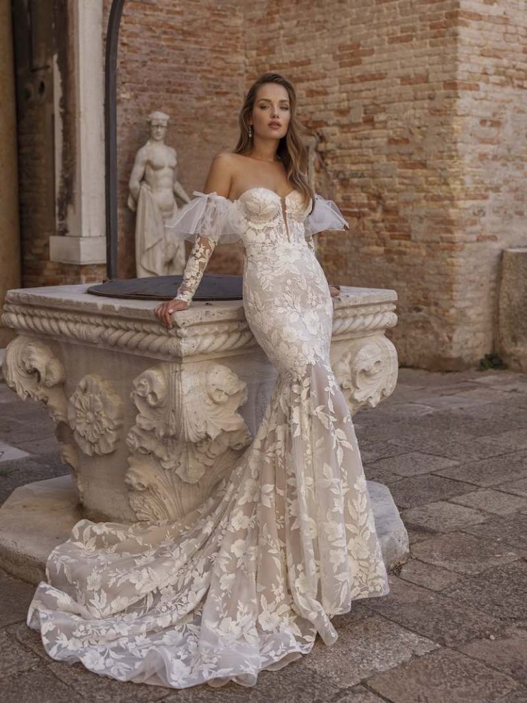 In questa foto una modella indossa un abito da sposa interamente ricamato. Le maniche lo sono altrettanto. Il vestito è un modello a semi sirena 2023, con bustier con scollatura a cuore