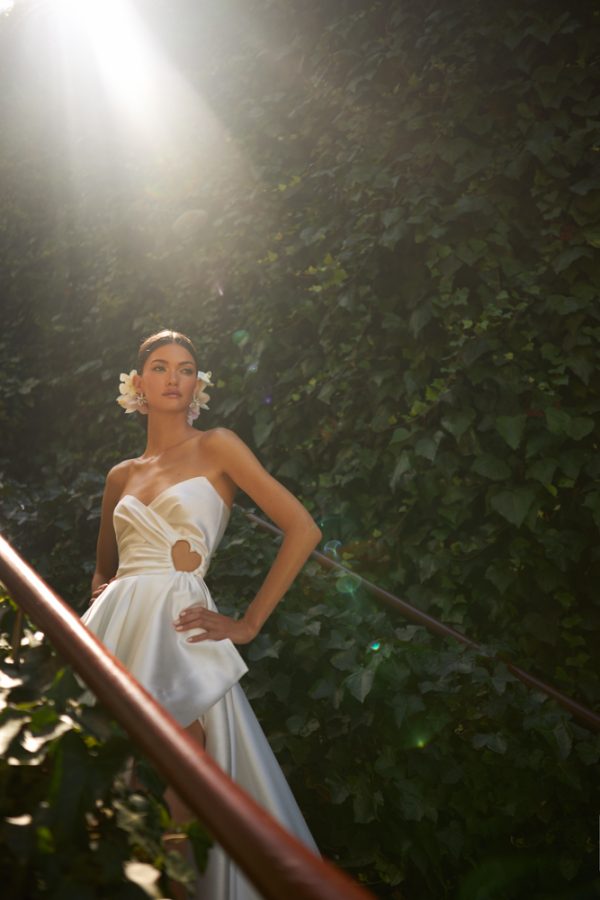 In questa foto una modella, su una scalinata, indossa un abito da sposa in mikado di seta, con scollo a cuore e un originalissimo taglio cut-out a forma di cuore. Il modello fa parte dei vestiti Inbal Dror 2023