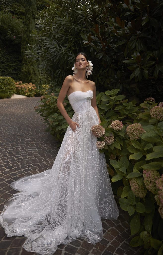 In questa foto una modella indossa un abito da sposa dalla lina ad A con bustier in tulle e gonna con ricami luminosi. Il modello è di Inbal Dror 2023