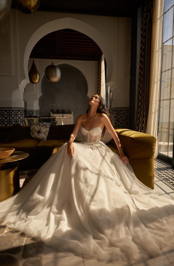 In questa foto una modella su un divano mentre indossa un abito da sposa con bustier e gonna scivolata e leggera della collezione Inbal Dror 2023