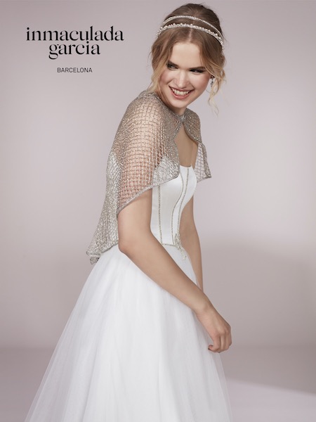 In questa foto una modella indossa una piccola mantella lurex su un abito da sposa Inmaculada Garcia 2023 