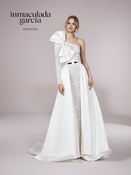 In questa foto una modella indossa uno splendido abito da sposa Inmaculada Garcia 2023: il vestito è monospalla, con un fiocco maxi applicato sulla spalla. L'abito è interamente in pizzo, con un sovragonna in mikado. Sia il fiocco che il sovragonna sono rimovibili