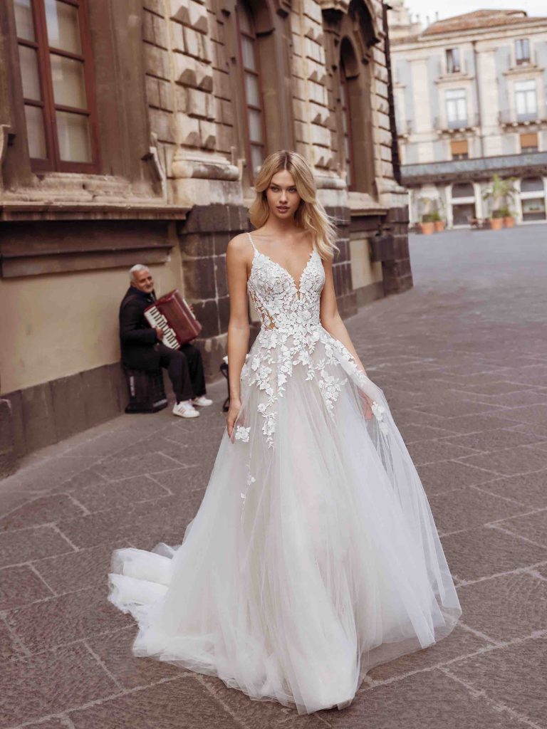 In questa foto una modella passeggia indossando un abito da sposa con top in pizzo floreale e gonna in tulle, della collezione sposa Modeca 2023
