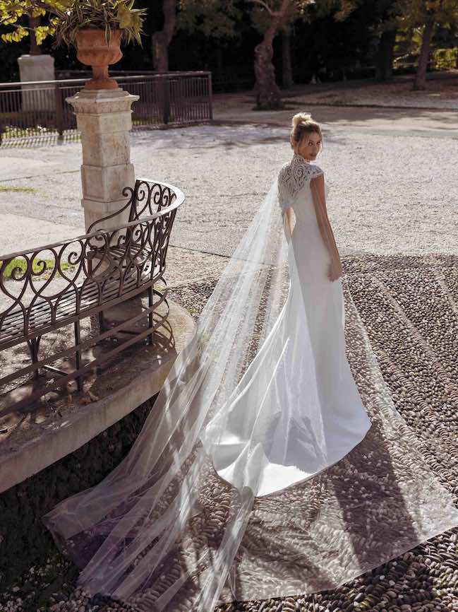 In questa foto una modella indossa un abito da sposa con lunga mantella in tulle di Modeca 2023