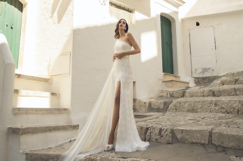 In questa foto una modella indossa un abito da sposa Pietro Demita Haute Couture 2023 monospalla, con gonna con frange e spacco. A cingere la vita, una cintura maxi in gold