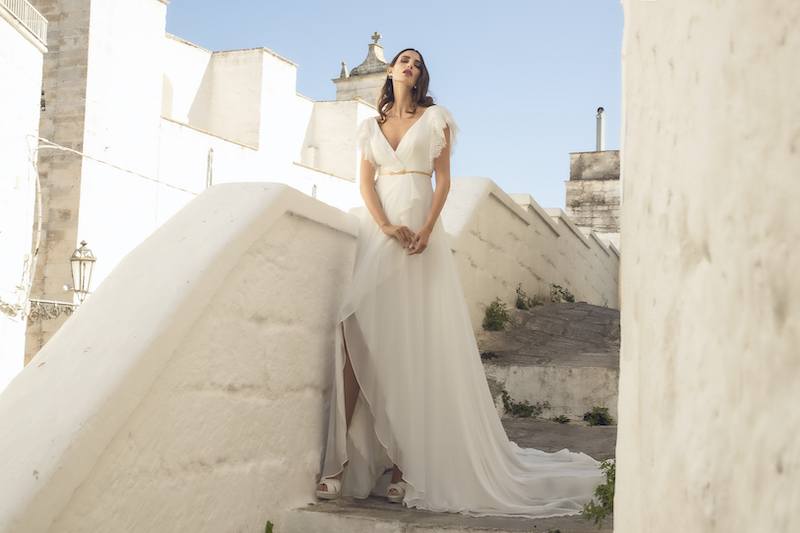 In questa foto una modella indossa un abito da sposa scivolato e leggero, tagliato sotto il seno e segnato da un cinturino dorato